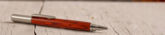 A handcrafted pen with a rich, reddish-brown wooden barrel featuring dark grain patterns, paired with polished metal accents. The pen rests diagonally on a weathered wooden surface, highlighting its artisanal detail and natural elegance.